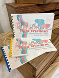 Three spiral-bound notebooks titled "Wild Wisdom: 12 Animal Adventures in Truth" are stacked on a rustic wooden surface with beige fabric around them. The covers feature colorful illustrations of animals including a giraffe, elephant, lion, and buffalo, with blue, red, pink, yellow, and blue spiral binding.