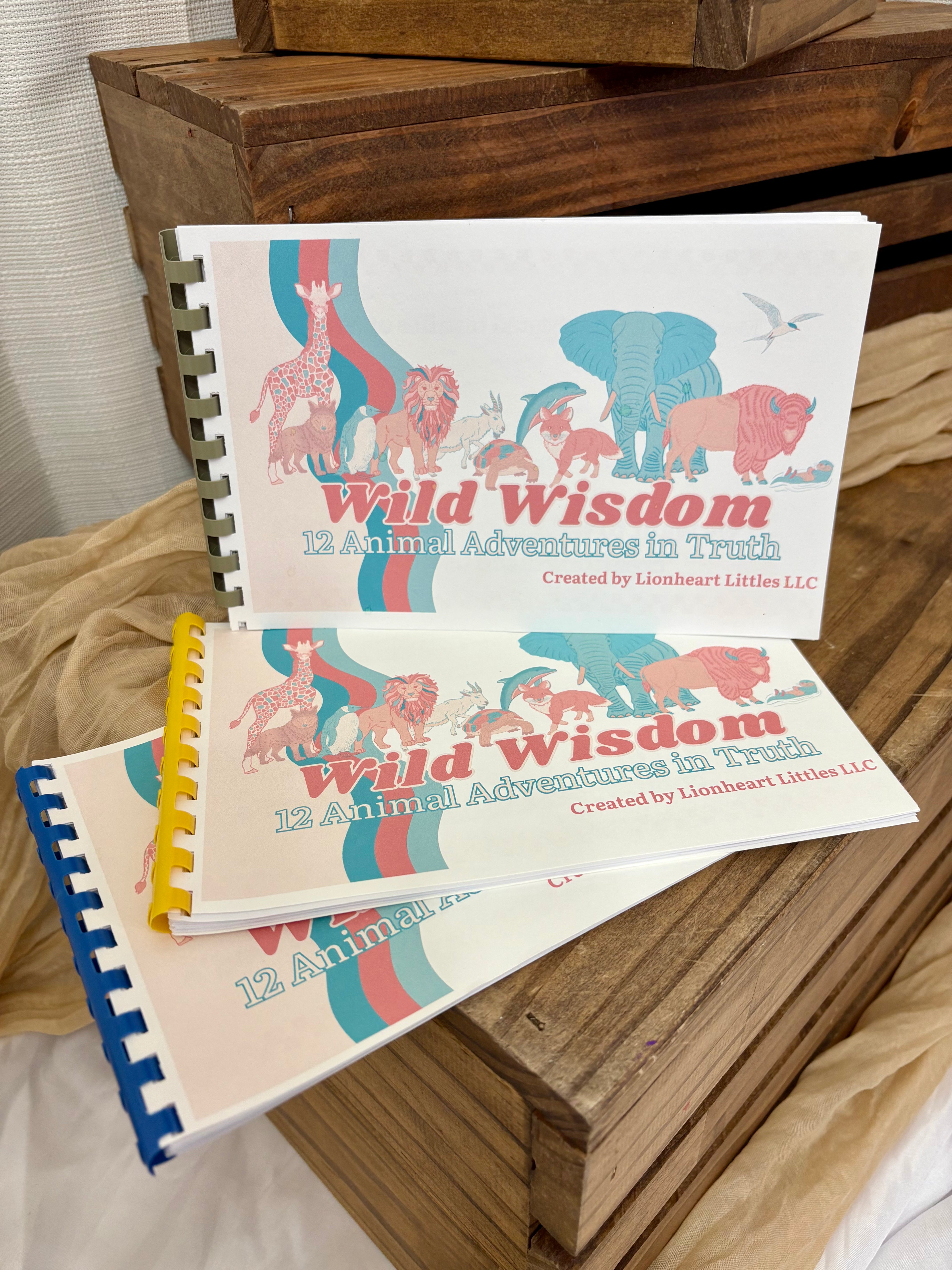 Three spiral-bound notebooks titled "Wild Wisdom: 12 Animal Adventures in Truth" are stacked on a rustic wooden surface with beige fabric around them. The covers feature colorful illustrations of animals including a giraffe, elephant, lion, and buffalo, with blue, red, pink, yellow, and blue spiral binding.