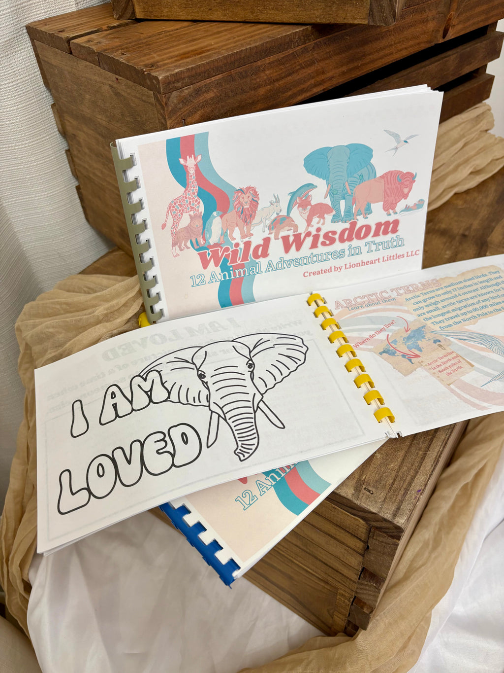 A spiral-bound children's book set displayed on rustic wooden crates and draped fabric. The front cover shows the title 'Wild Wisdom 12 Animal Adventures in Truth' with colorful animal illustrations, and the open pages show a black-and-white elephant coloring page with the words 'I AM LOVED' and more colorful interior pages.